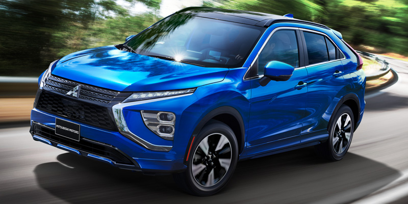 Front-three-quarter view of a vibrant blue Mitsubishi Eclipse Cross driving on a winding road with a motion-blurred forest background.