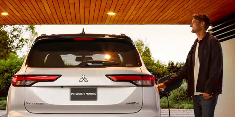 Rear view of a silver Mitsubishi Outlander PHEV being plugged in by a smiling man under a wood-paneled carport.