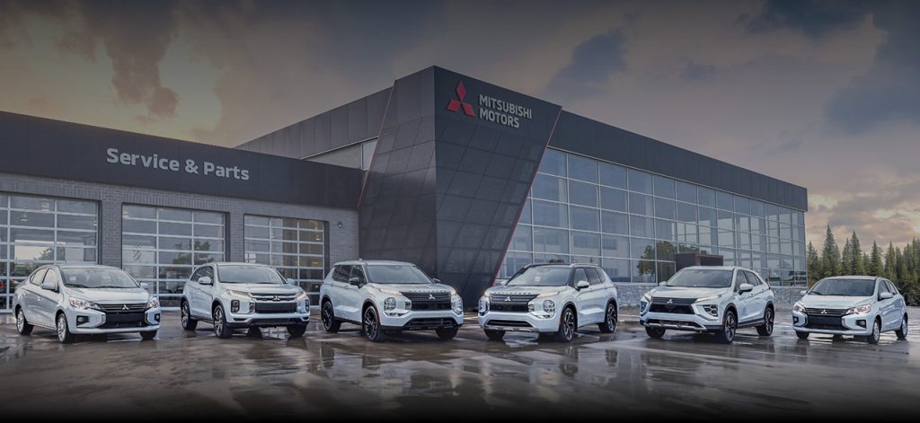 Six white Mitsubishi vehicles parked side-by-side on a wet asphalt surface in front of a modern Mitsubishi Motors dealership with a "Service & Parts" wing under a cloudy evening sky.