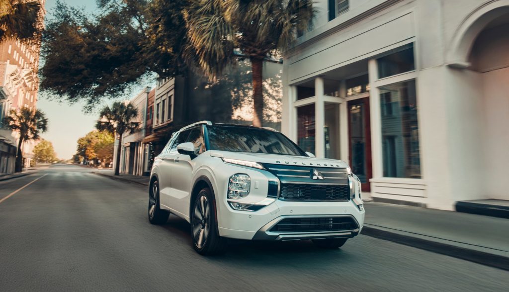 Front-three-quarter view of a white Mitsubishi Outlander driving down an empty city street lined with palm trees and historic buildings.