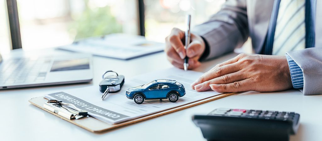 Close-up of a person filling out a car contract agreement next to a tiny toy car, key, and calculator.