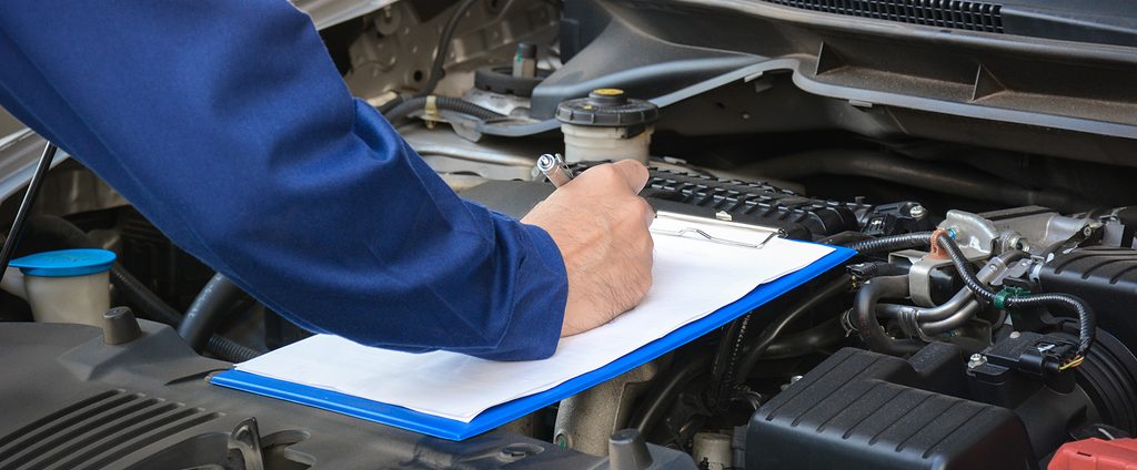 Close-up of a mechanic in a blue uniform writing on a clipboard while inspecting an open car engine bay.