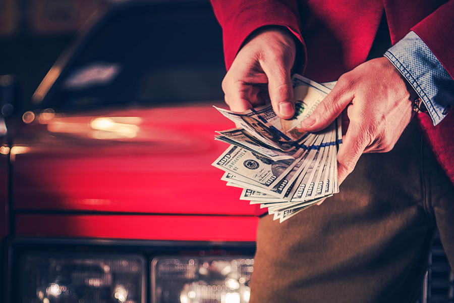 Close-up of a person counting a fan of US dollar bills in front of a red vehicle.