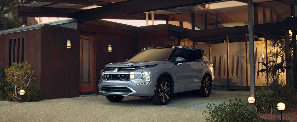 A light gray Mitsubishi Outlander SUV parked in the carport of a mid-century modern home at dusk.
