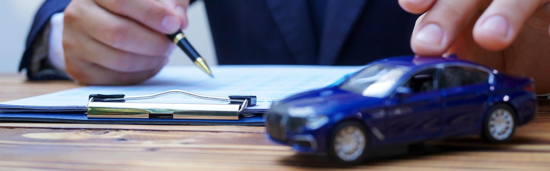 A person filling out contract paperwork at a desk next to a blue miniature toy car, representing auto insurance, loan, or sales agreements.